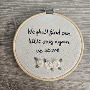 A 4 inch light wood embroidery hoop with linen fabric. Words "We shall find out little ones again up above" embroidered in black with 3 cream roses underneath.