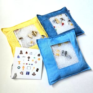 Three I spy bags shown, one with the image card displayed. One bag is dark blue, one lighter blue and one yellow. The bags are filled with Catholic symbols of our faith